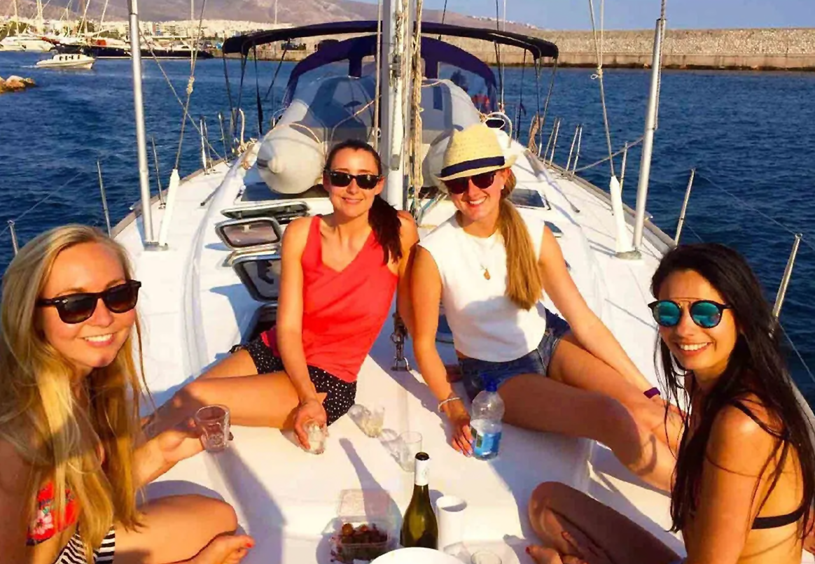 Four women on a sailboat deck, smiling in sunglasses, holding drinks, sunny day.