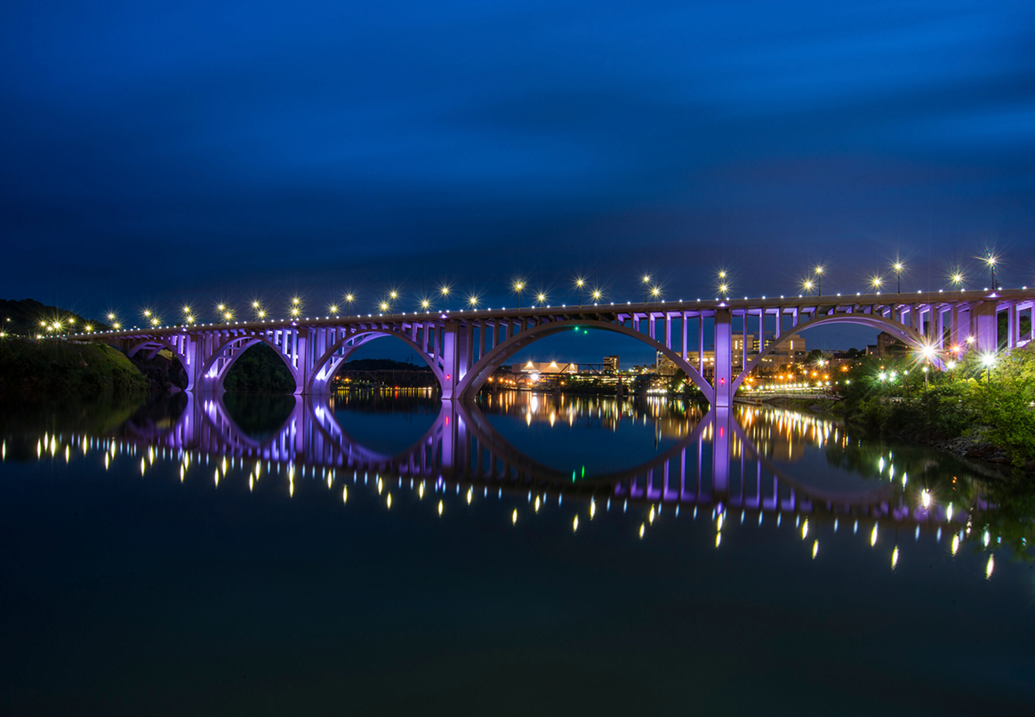 Night view of Knoxville river bank a purple-lit bridge reflecting in calm water; city lights in the background.