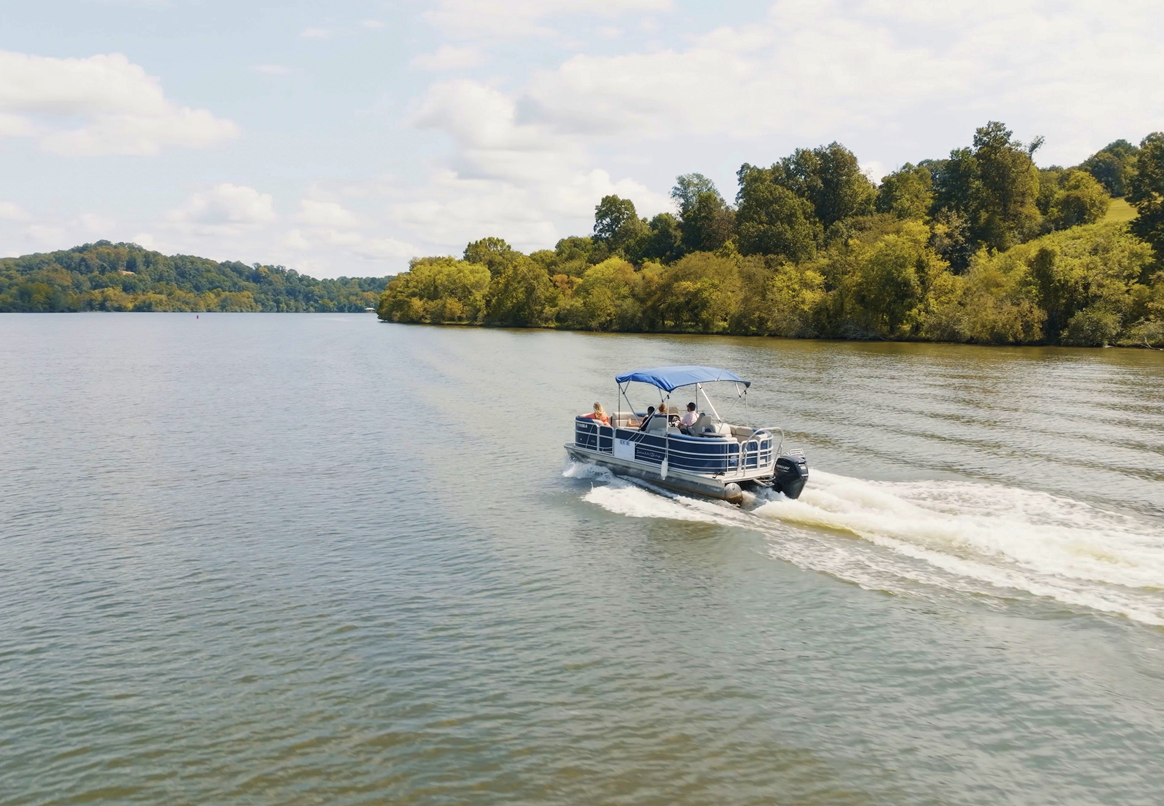 A pontoon boat cruises across a lake. Green trees line the shore under a partly cloudy sky.