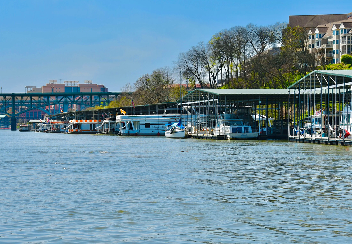 Boats docked on a river under covered slips. A green bridge and buildings are in the background. Blue water and sky.