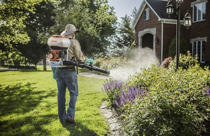 A man is spraying plants in a yard with a backpack sprayer.