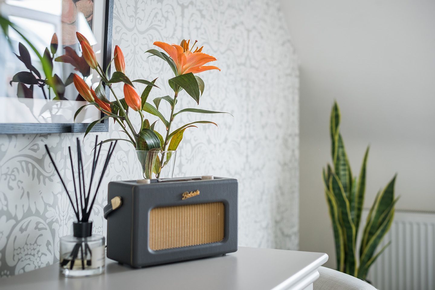 Grey bedroom, calm vibe, with Farrow & Ball wallpaper, orange lilies in glass vase and grey Roberts radio