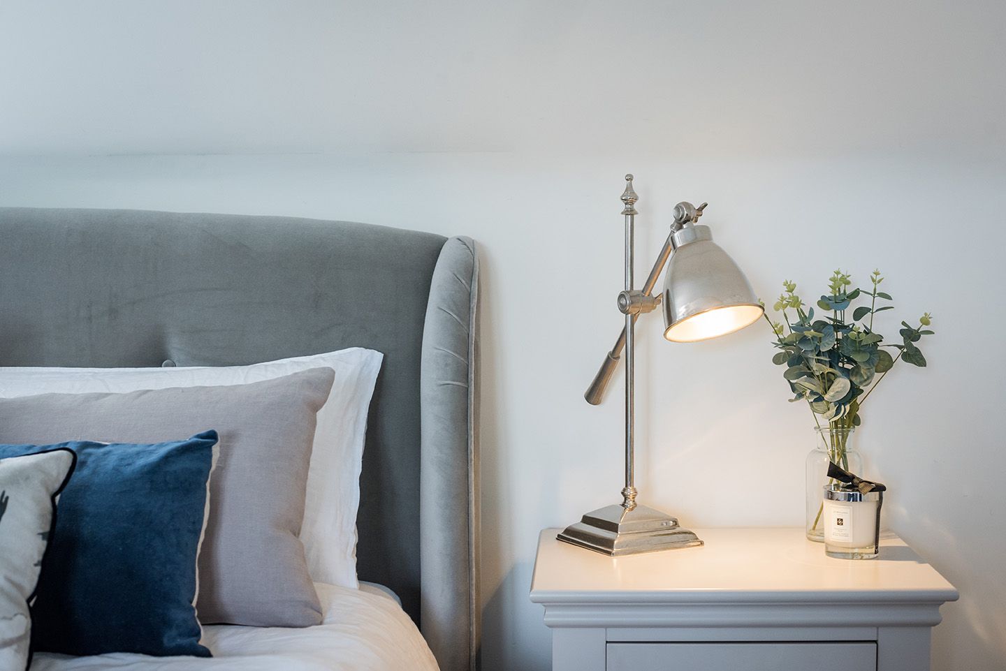 Grey velvet bed with bedside table, chrome table lamp and cushions, soft calm tones