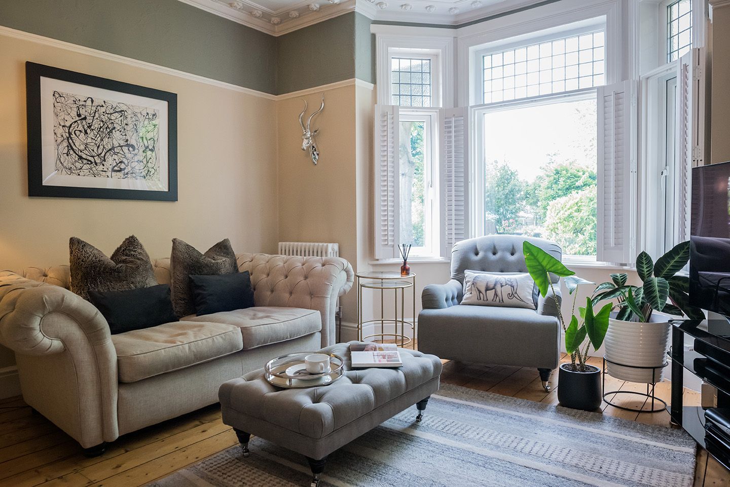 Cream and green living room with cream chesterfield sofa and buttoned footstool. Bay window with shutters and plants