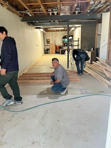Men working on a construction project inside a room. One squats near wooden beams, while two others work nearby.