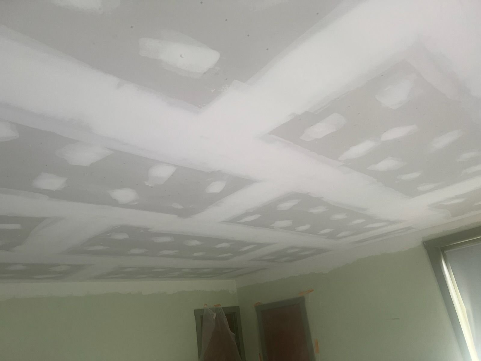 Ceiling with drywall squares, taped joints, and spackling. Light green walls, window, and doors in the room.