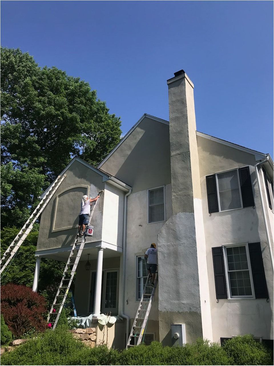 Two people on ladders painting a house exterior on a sunny day.