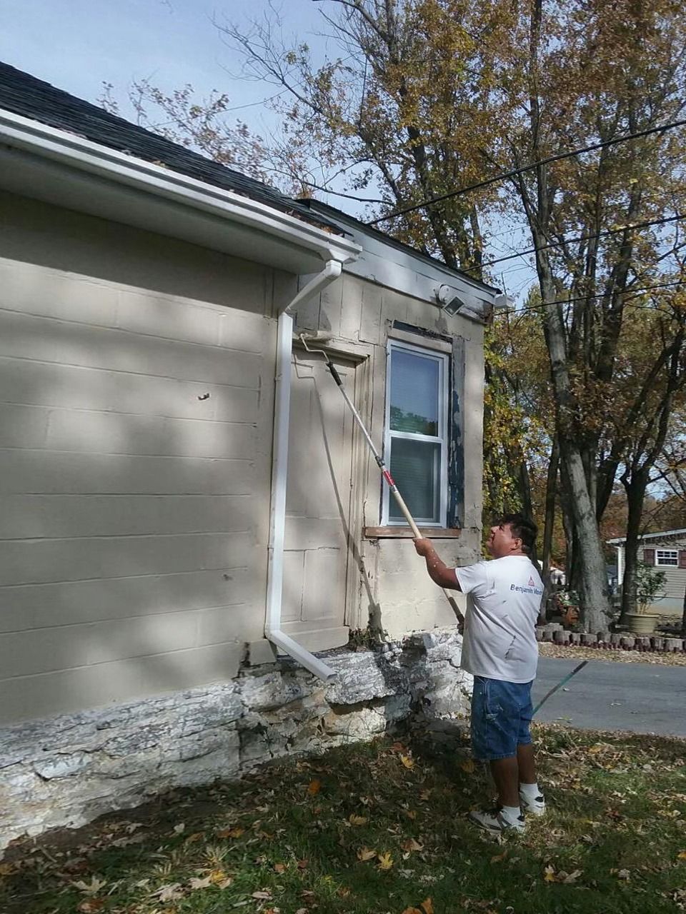 Man paints house exterior with long-handled brush. Beige wall, white trim, blue sky, and fall trees.