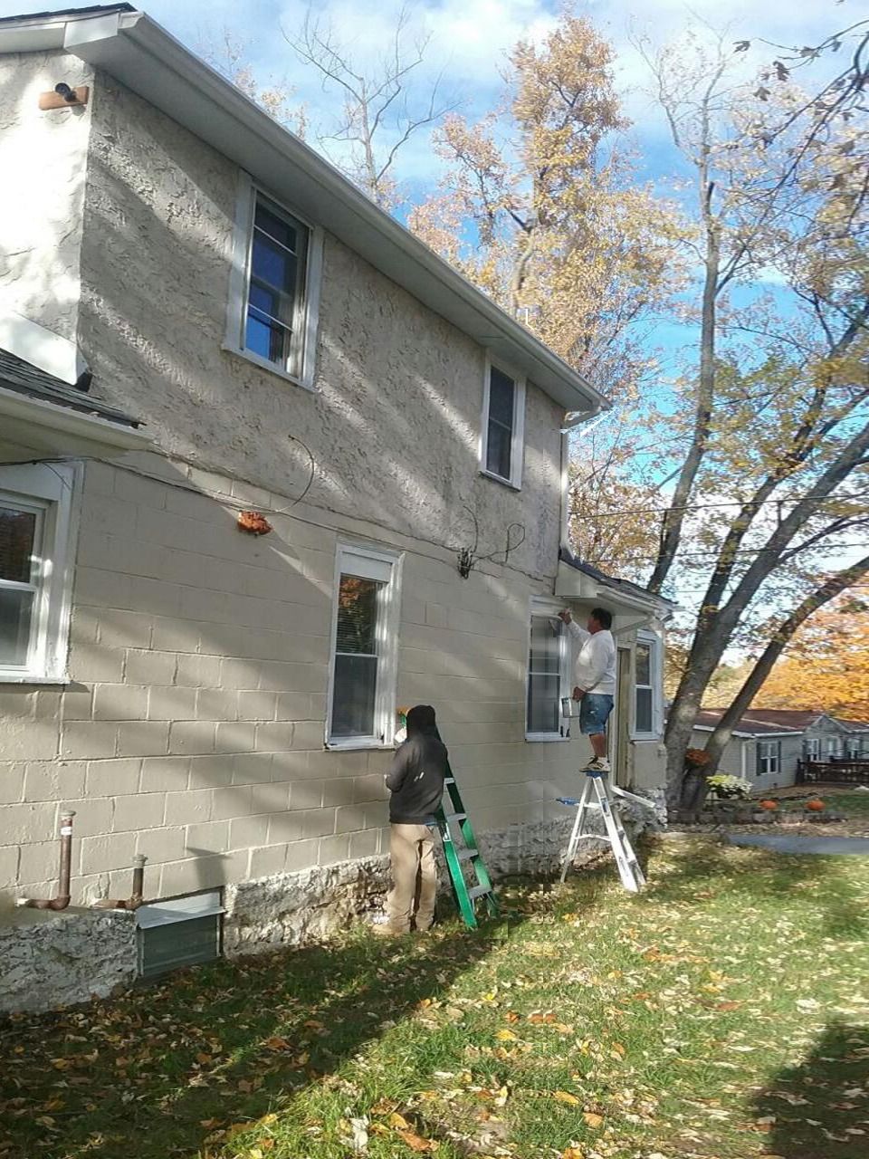 Two people on ladders working on the side of a two-story house with trees and fall foliage.