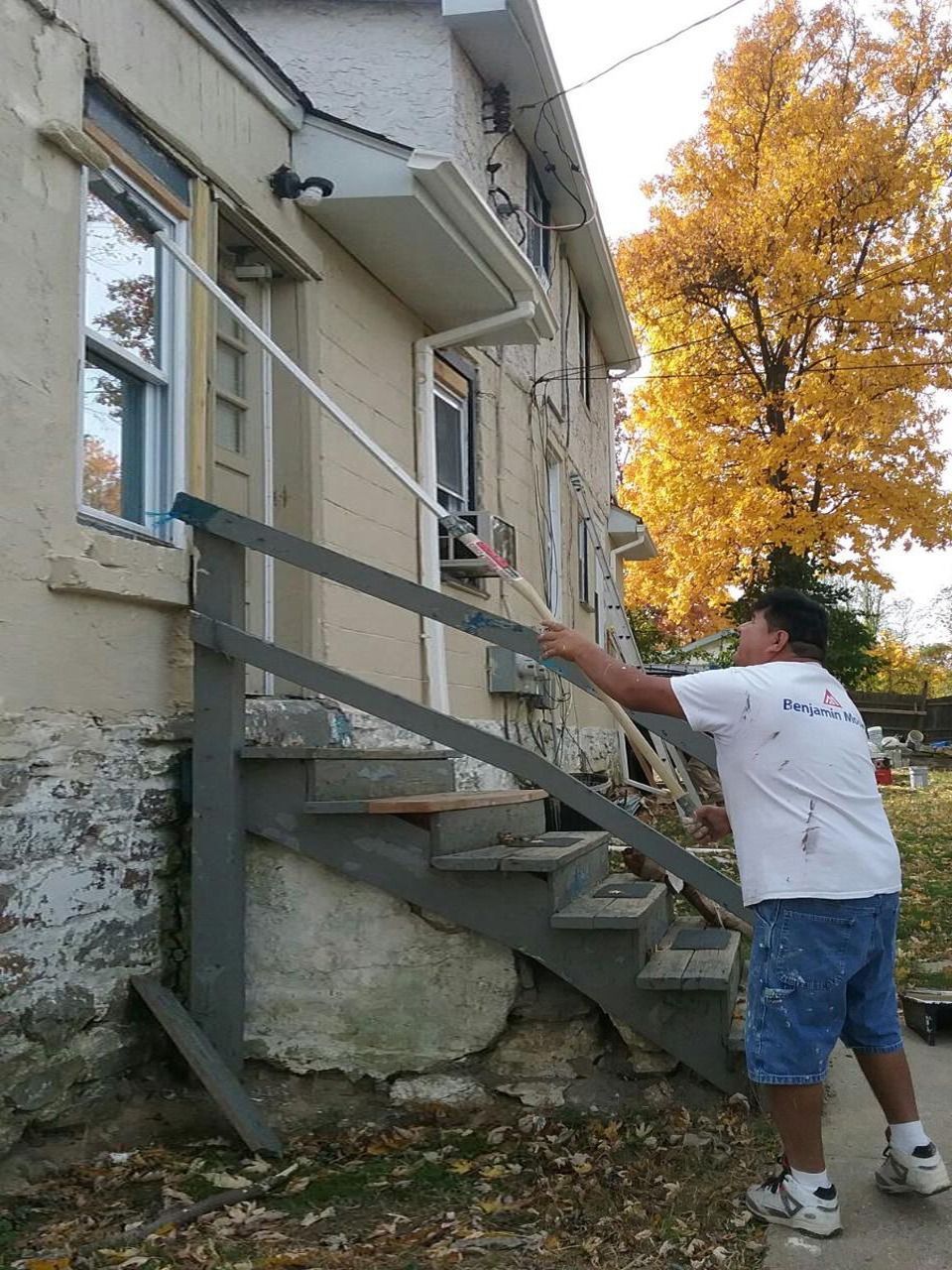 Man painting a window from a wooden staircase, outside of a light-colored building.