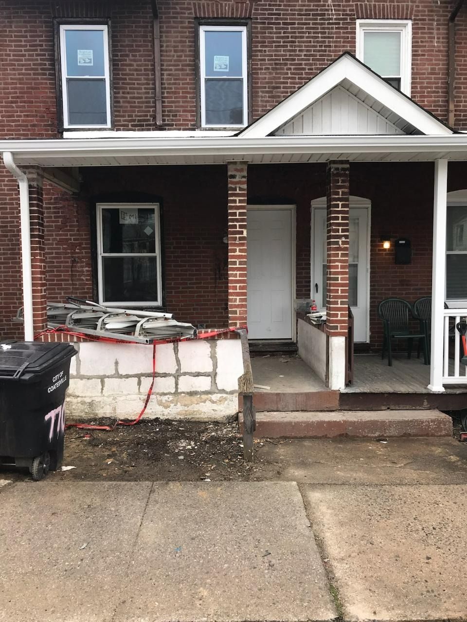 Brick home exterior with porch undergoing renovation; front door, windows, trash bin, and sidewalk visible.