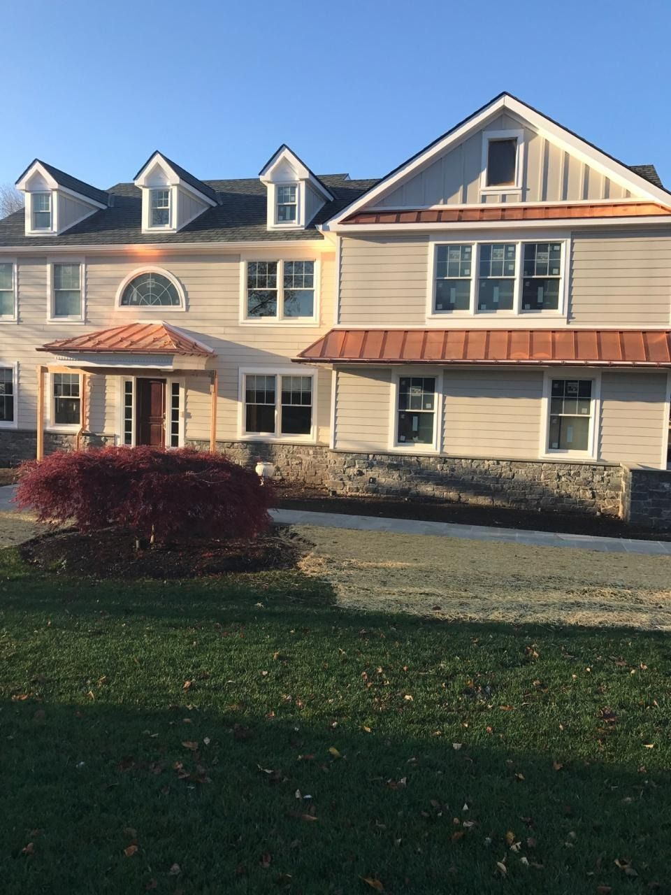 Two-story house with light gray siding, copper roof accents, stone foundation, and small red bush in the yard.
