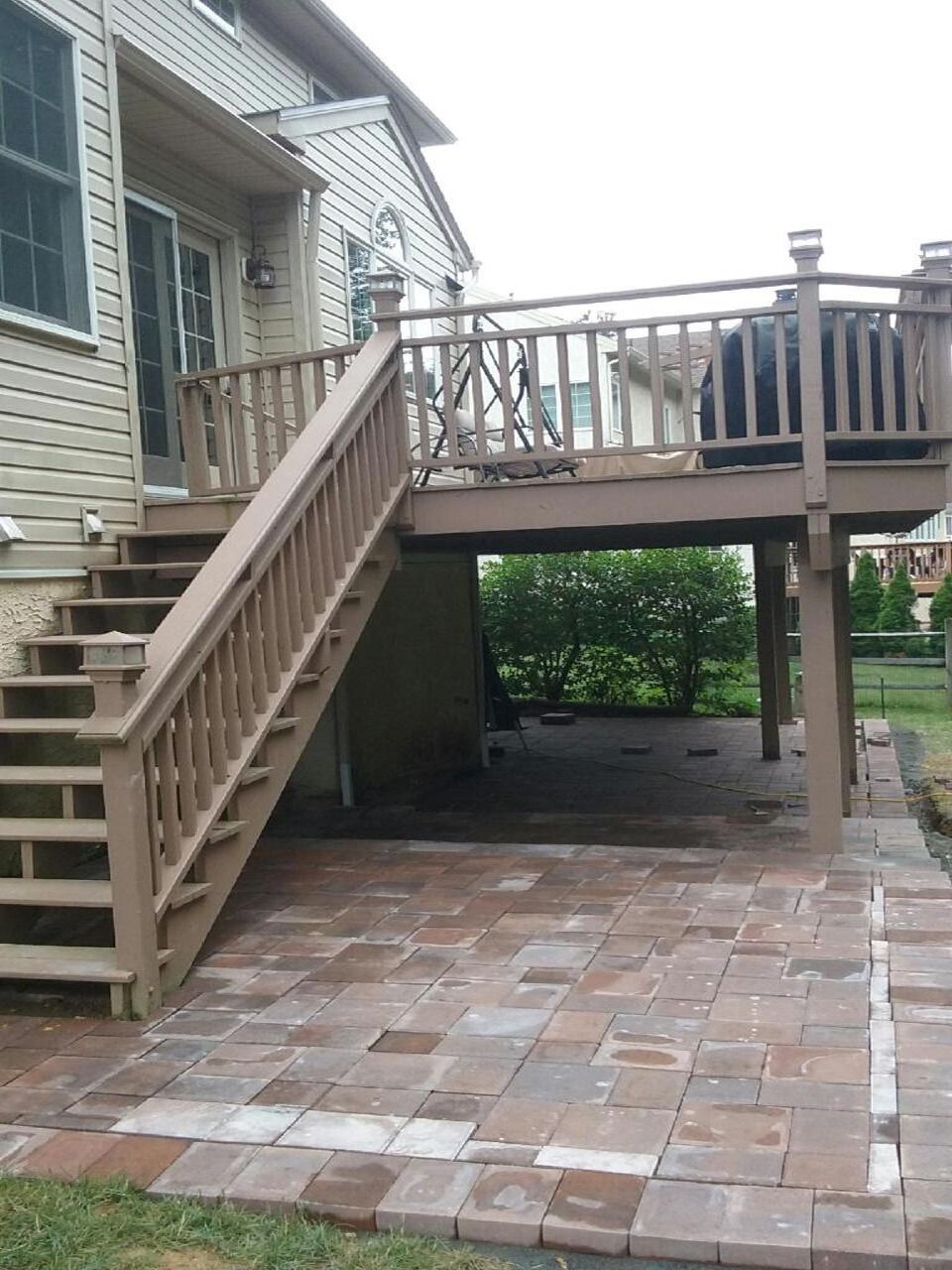 Brown wooden deck with stairs leading to a brick patio, beneath the deck is a shaded area.