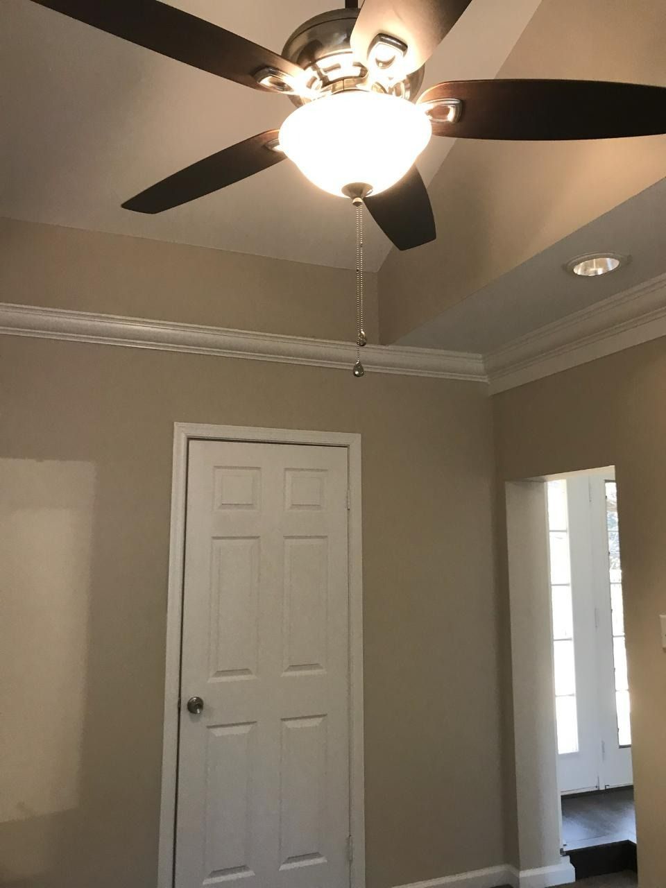 A room with a ceiling fan and a closed white door. Beige walls, white trim.