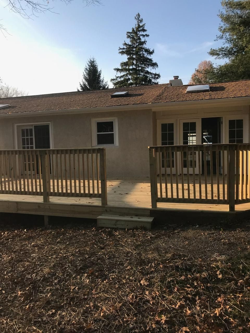 Wooden deck attached to a light-colored house, surrounded by brown leaves and trees under a blue sky.