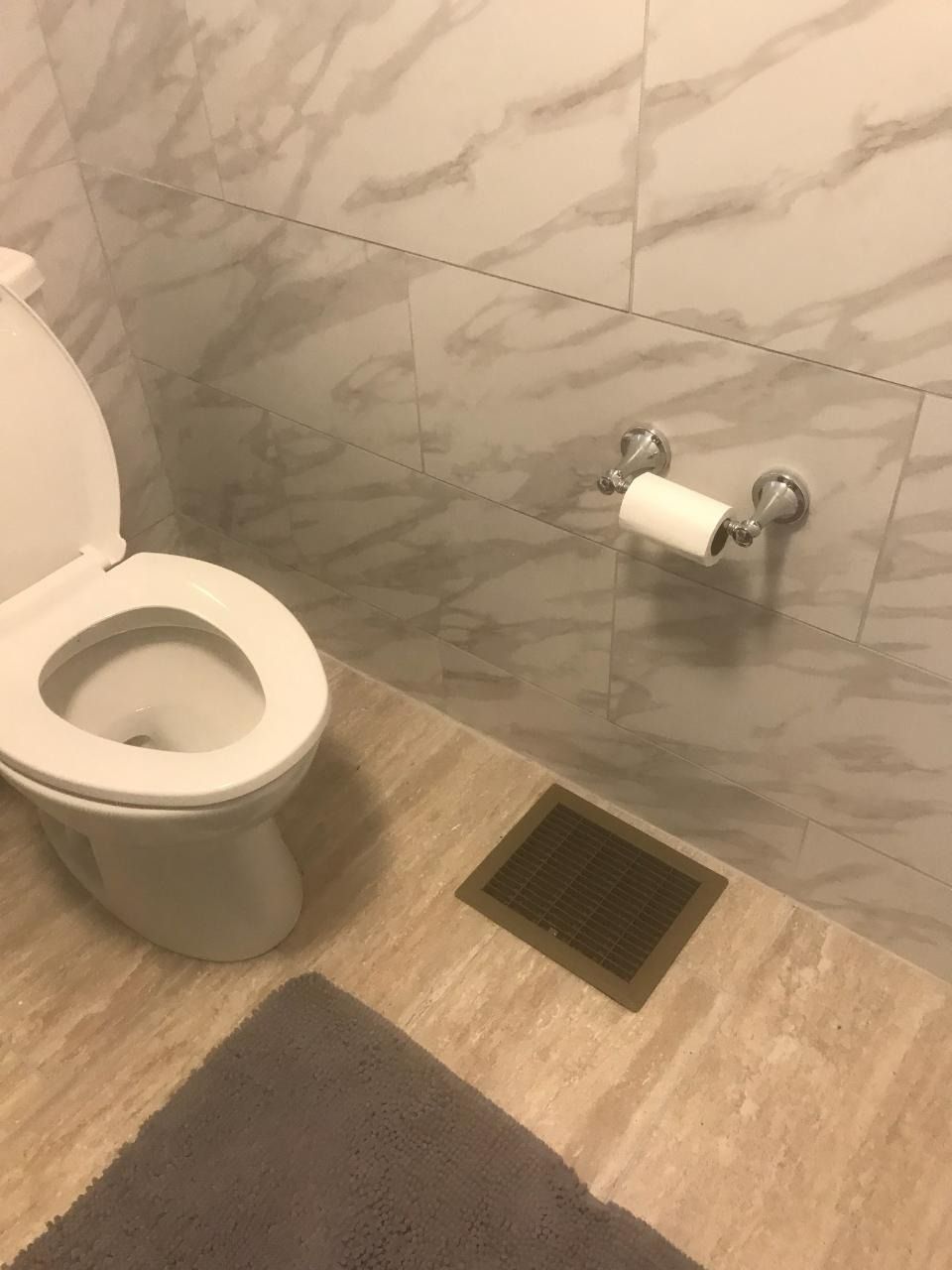 Toilet with seat up, gray bath mat, and toilet paper holder on marble tiled wall.