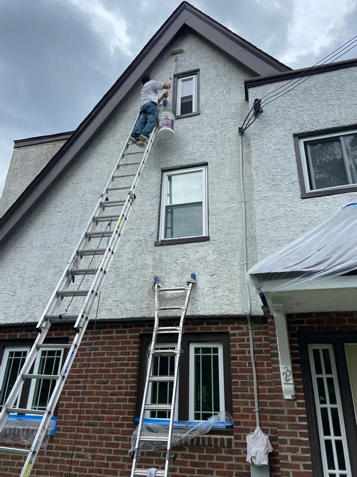 Person painting the exterior of a house from a tall ladder. Cloudy sky, white and brick walls, brown trim.