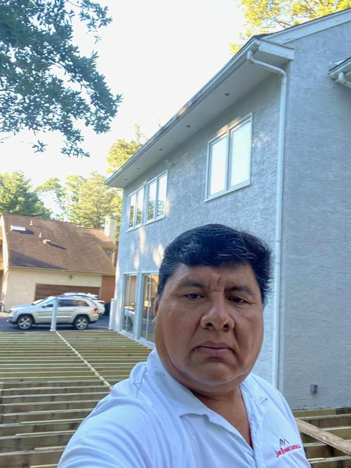 Man on unfinished deck with house in background, car parked, and cloudy sky.