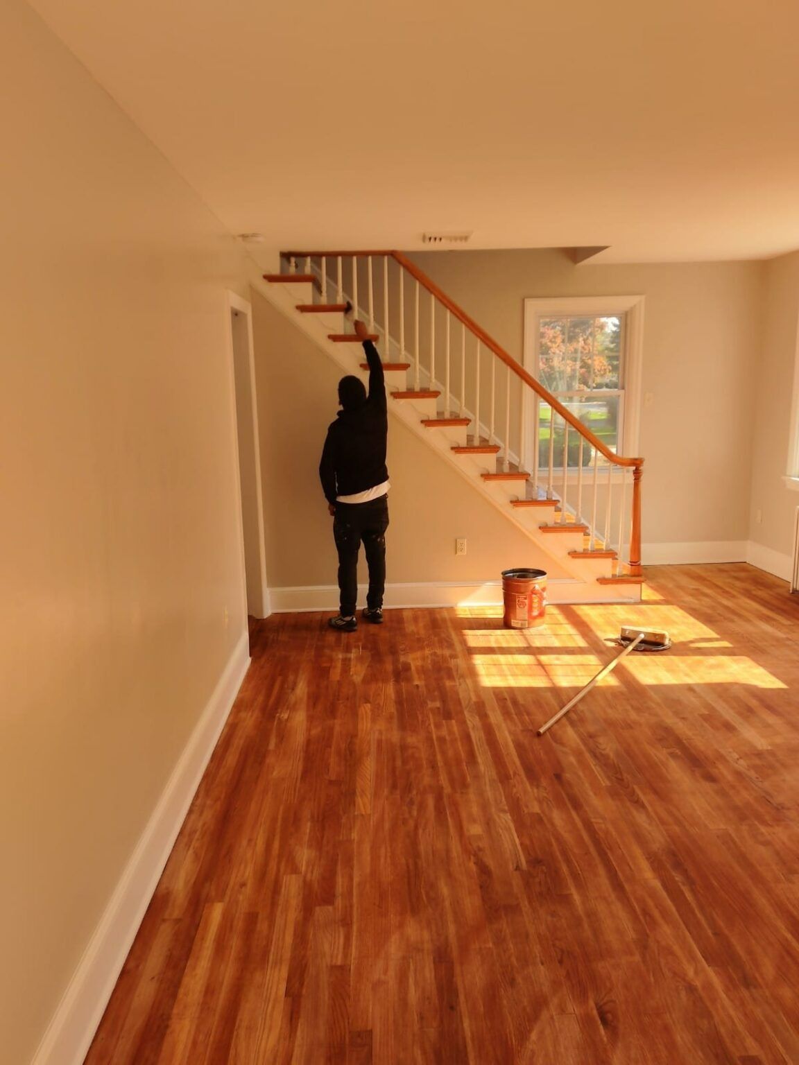 Person painting a staircase in a room with hardwood floors. Sunlight streams in the window.