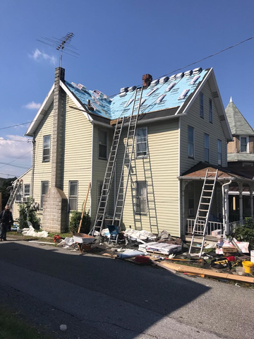 House with roof partially covered with blue underlayment; several ladders and debris on the ground, roof repair in progress.