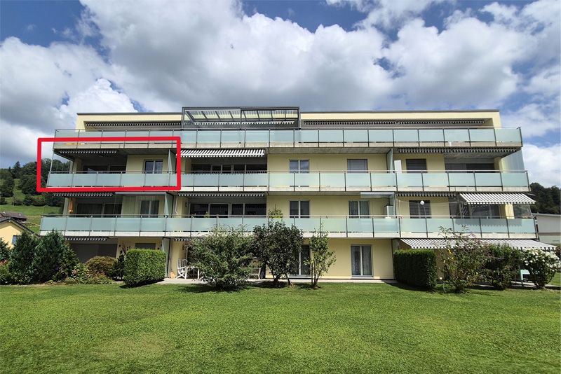 Ein gelbes dreistöckiges Wohnhaus mit Balkonen und einem roten Kasten, der eine Wohnung im obersten Stockwerk hervorhebt, steht auf einem grünen Rasen.