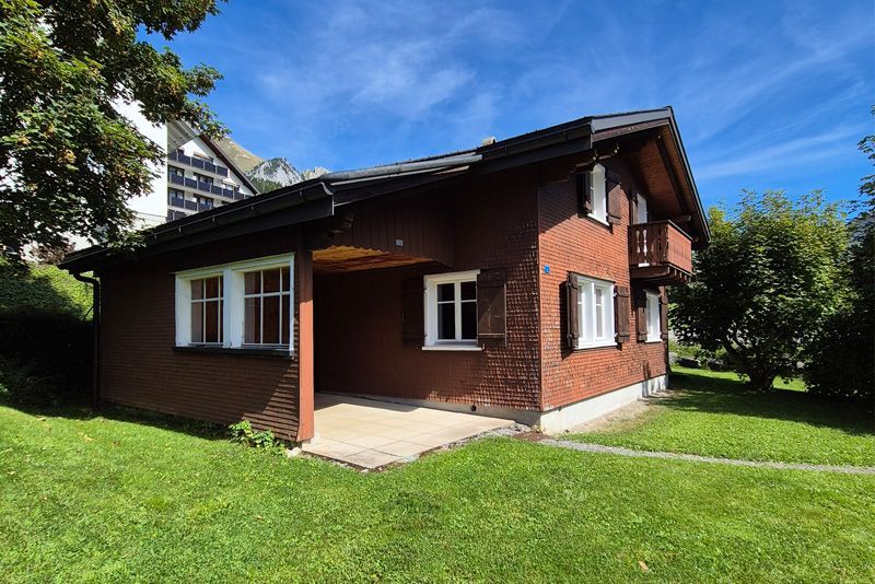 Ein zweistöckiges braunes Chalet mit weiß gerahmten Fenstern, einer überdachten Terrasse und einem Holzbalkon, eingebettet in einen Rasengarten.