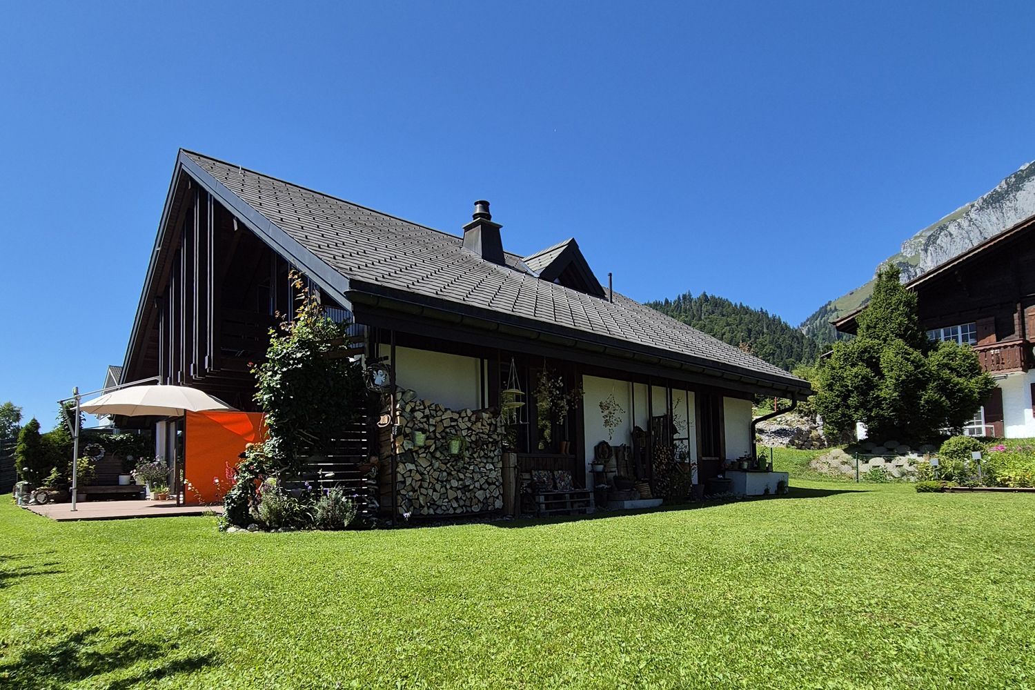 Ein zweistöckiges braunes Chalet mit weiß gerahmten Fenstern, einer überdachten Terrasse und einem Holzbalkon, eingebettet in einen Rasengarten.