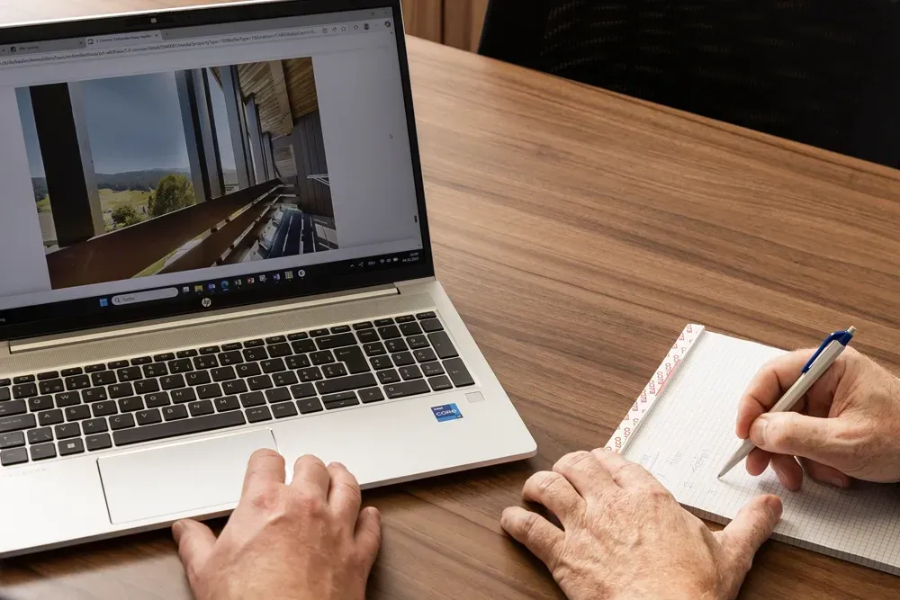 Eine Person benutzt einen Laptop, auf dem eine Innenraumdarstellung angezeigt wird, während sie sich Notizen auf einem Block auf einem Holzschreibtisch macht.