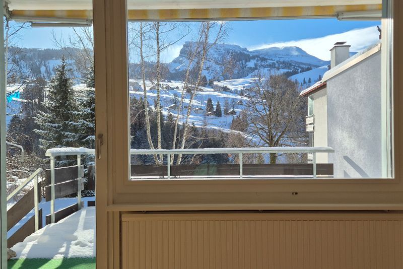 Blick aus einem Fenster auf eine schneebedeckte Berglandschaft mit Bäumen, einem Balkon und klarem blauen Himmel.
