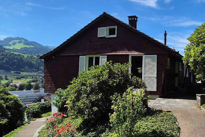 Ein braunes Holzhaus mit weißen Fensterläden, eingebettet in einen Hang, mit einem üppigen Garten und Bergblick im Hintergrund.