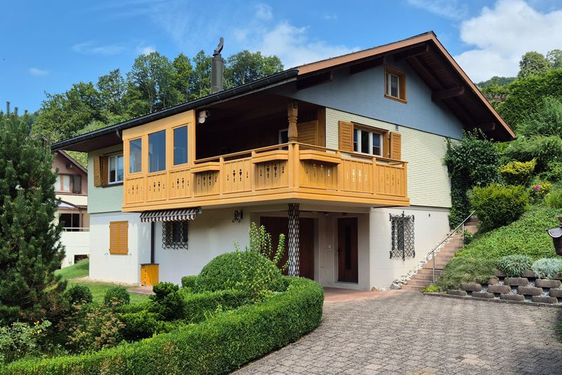 Ein zweistöckiges Haus mit einem markanten Holzbalkon, blau-beiger Fassadenverkleidung, umgeben von gepflegten Hecken und einer Auffahrt.