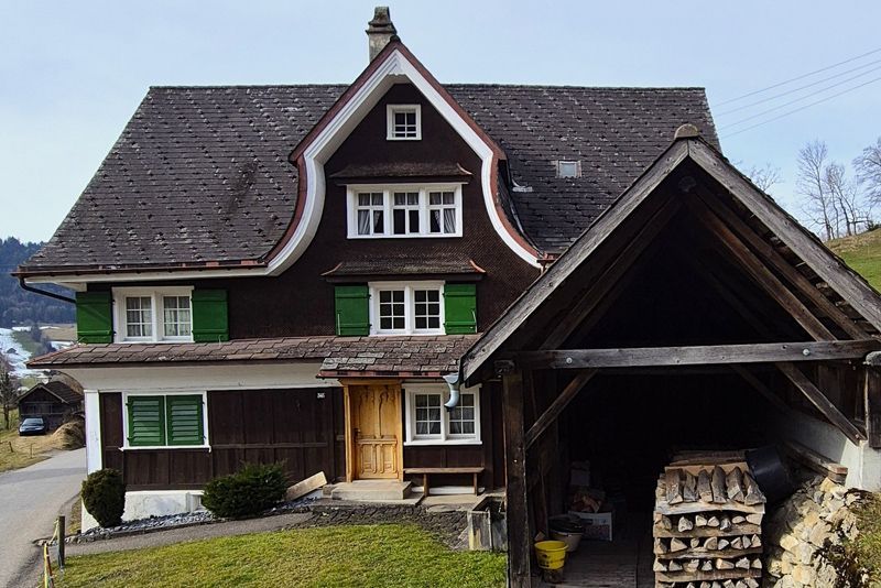 Ein traditionelles dunkles Holzhaus mit grünen Fensterläden und einem angebauten offenen Holzunterstand vor ländlicher Kulisse.