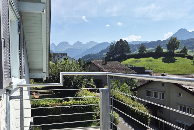 Ein Balkon mit Blick auf ein üppig grünes Tal mit Häusern, Bäumen und fernen Bergen unter blauem Himmel.