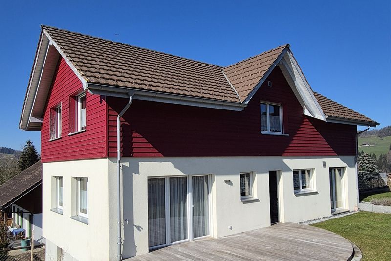 Ein zweistöckiges Haus mit weißem Stuck im Erdgeschoss, roter horizontaler Holzverkleidung im Obergeschoss und einem braunen Schindeldach.