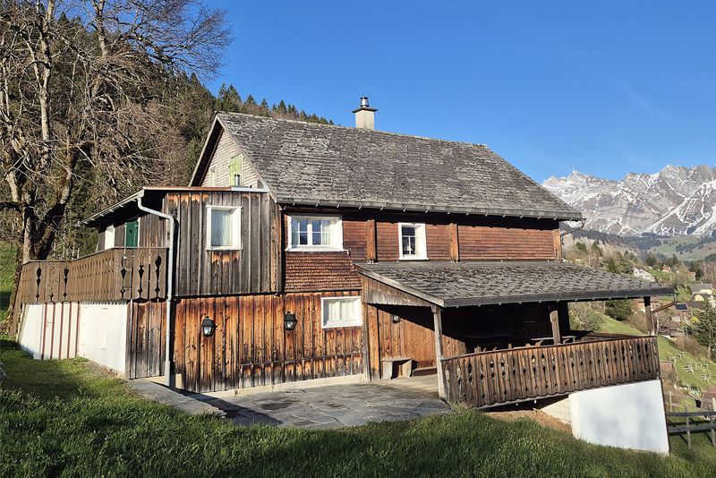 Ein rustikales Holzhaus mit Schindeldach liegt an einem grasbewachsenen Hang vor schneebedeckten Bergen unter klarem, blauem Himmel.