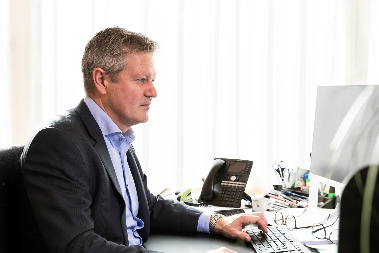 Eine Person arbeitet an einem Schreibtisch mit zwei Computermonitoren, tippt auf einer Tastatur, Bürounterlagen und ein Telefon befinden sich in der Nähe.