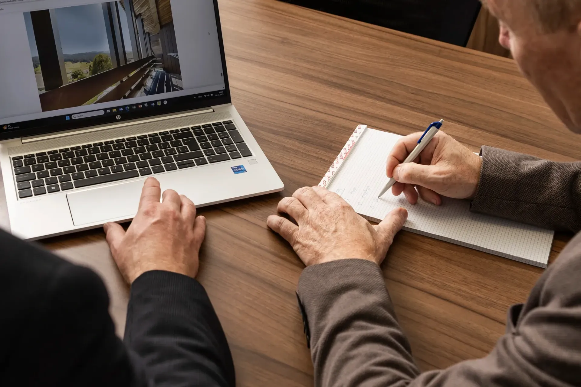 Zwei Personen arbeiten zusammen an einem Schreibtisch: Eine Person nutzt einen Laptop, auf dem Architekturpläne angezeigt werden, während die andere sich Notizen macht.