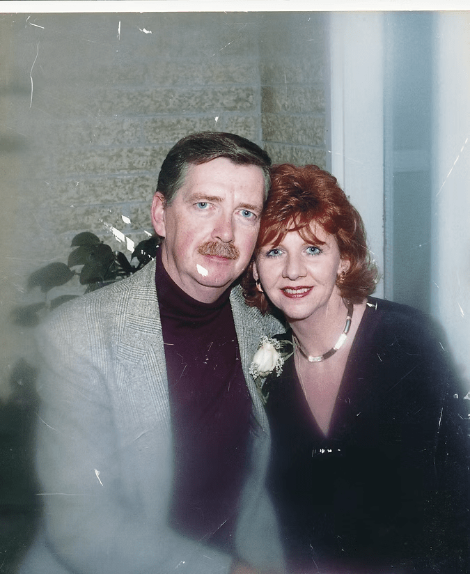 Two adults posing closely indoors, smiling, with a white flower on the man's jacket.