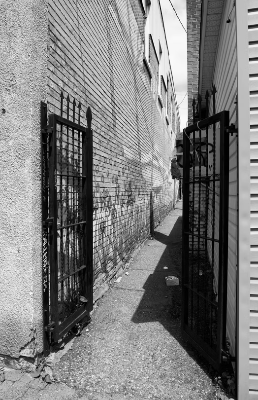 Narrow alley between brick buildings with black metal gates casting shadows on the ground.