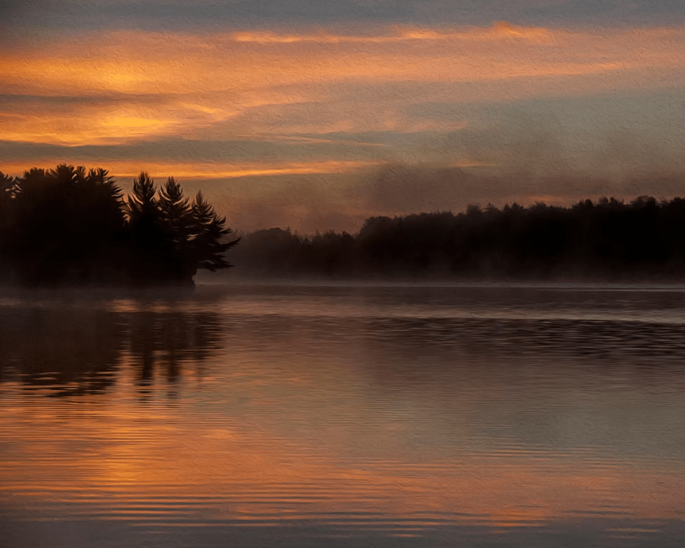 Misty lake at sunrise with orange sky and dark tree silhouettes reflected on calm water