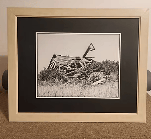 Framed black-and-white photo of a collapsed wooden structure in tall grass