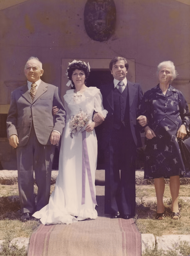Four people standing indoors at a formal wedding, with the bride in a white gown holding flowers.
