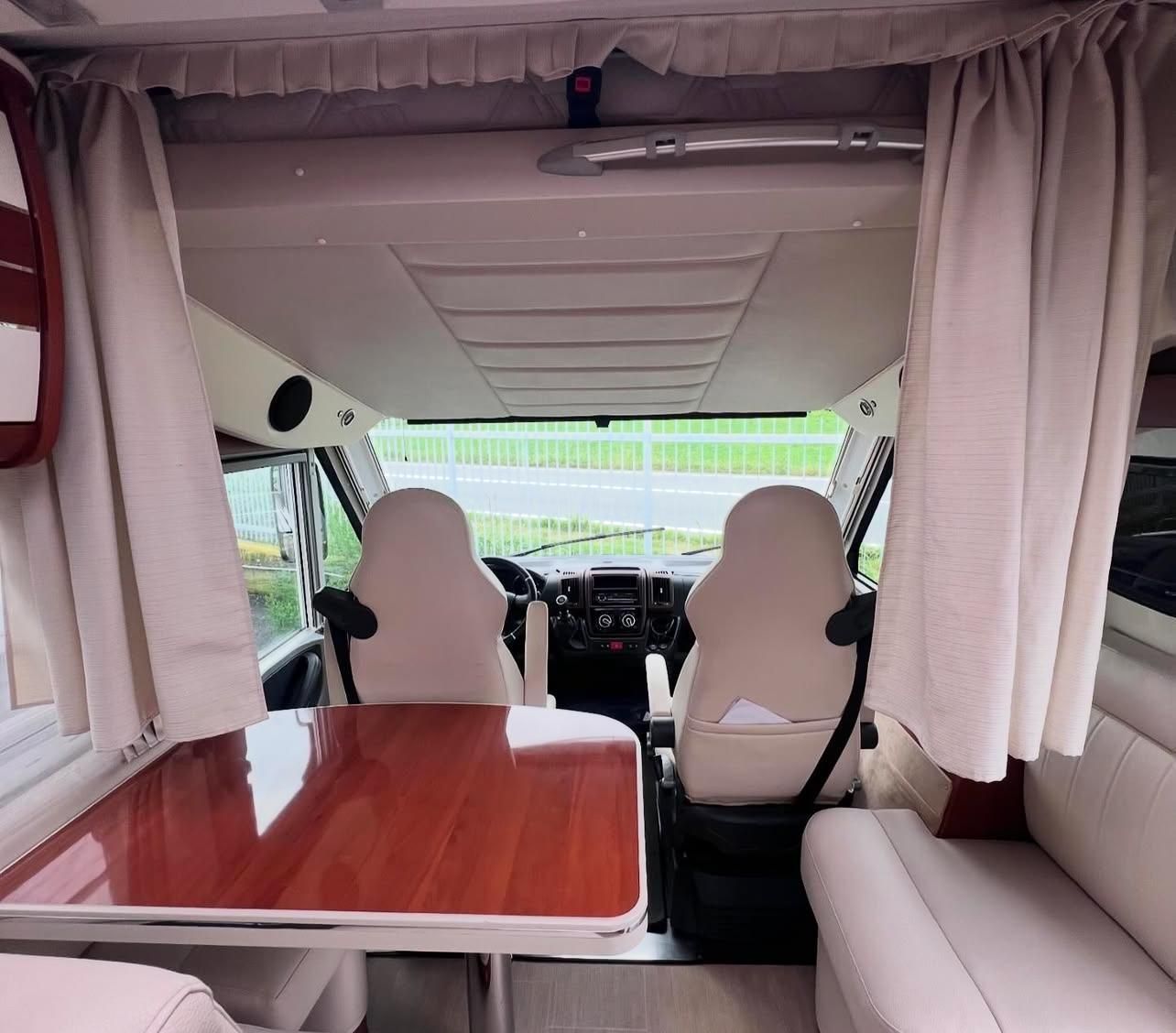 Interno di un camper con sedili anteriori, tavolo e tende; tonalità beige e legno.