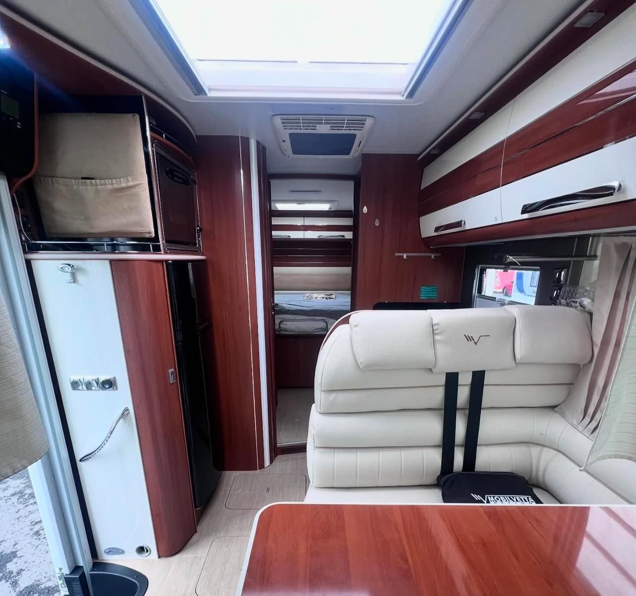 Vista interna di un moderno camper con mobili in legno bianco e scuro, una zona salotto e un lucernario.