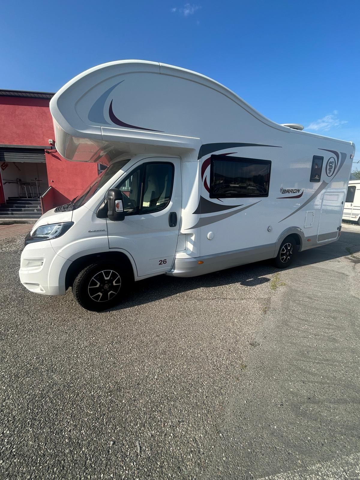 Camper bianco parcheggiato sulla ghiaia, contro un edificio rosso e un cielo blu.