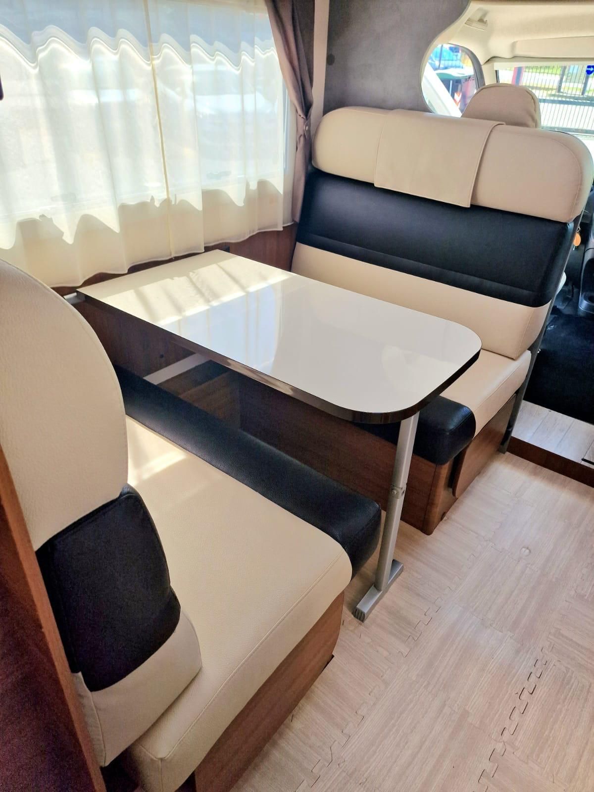 Dinette per camper con tavolo bianco, sedute color cuoio e nere e una finestra con tende trasparenti.