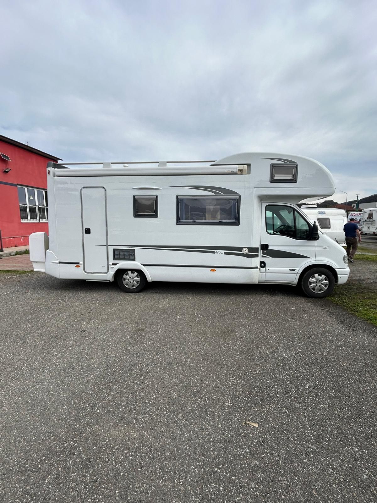 Camper bianco parcheggiato sulla ghiaia, sotto un cielo nuvoloso, vicino a un edificio rosso.