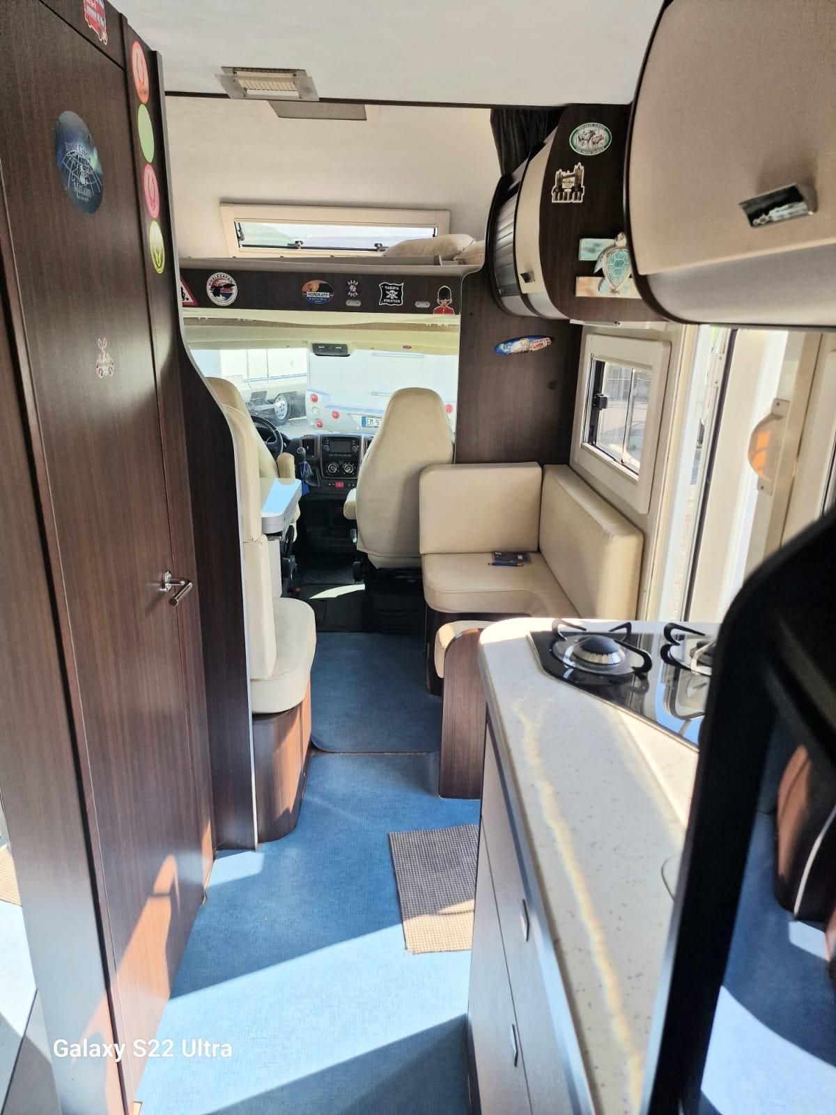 Interno di un camper: mobili marroni, sedili beige, pavimento blu e vista dei sedili anteriori.