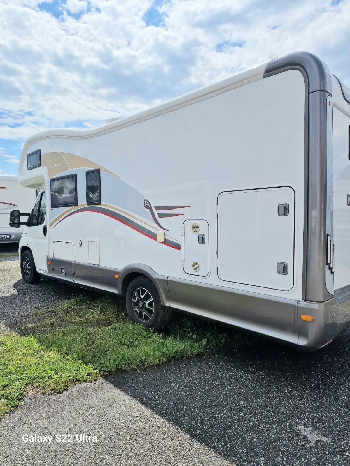 Veicolo ricreativo bianco e grigio con striscia decorativa rossa e oro parcheggiato sull'erba sotto un cielo nuvoloso.