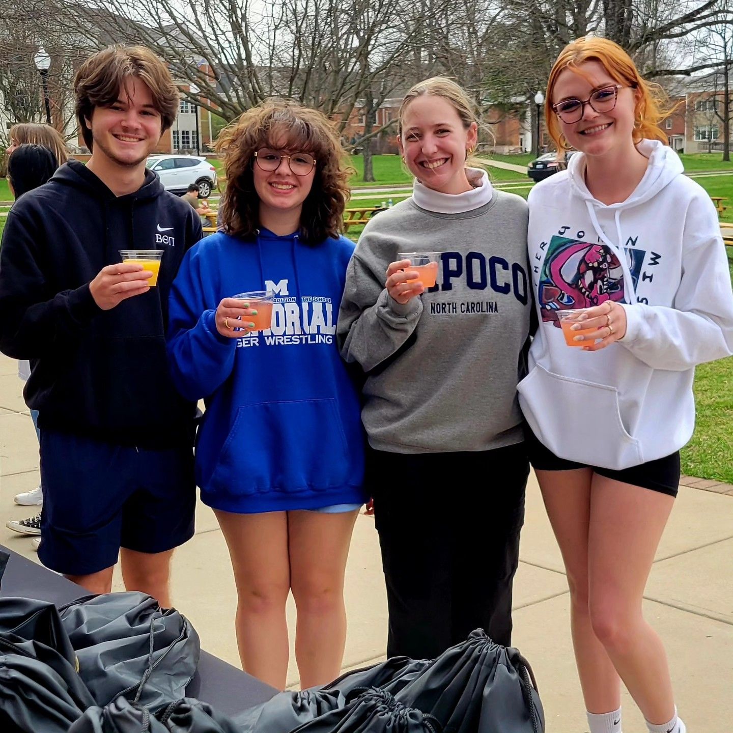 Four people smiling and holding drinks outdoors, wearing casual sweatshirts and standing on a sidewalk.